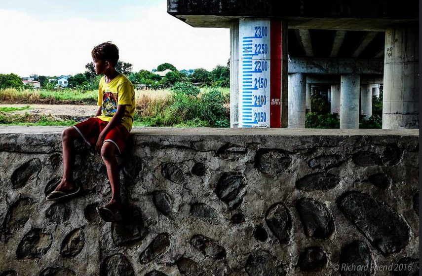 A young boy sits on top of a flood protection wall in Metro Manila. An informal community is settled under this bridge in a part of the river that is especially vulnerable to floods. Flood markers have been put in place to provide additional warning to the community - so that they can move to higher ground in case of an emergency. But the core of the problem is of lack of access to viable housing, employment and services that drives people to live in such vulnerable locations. Photo: © Richard Friend 