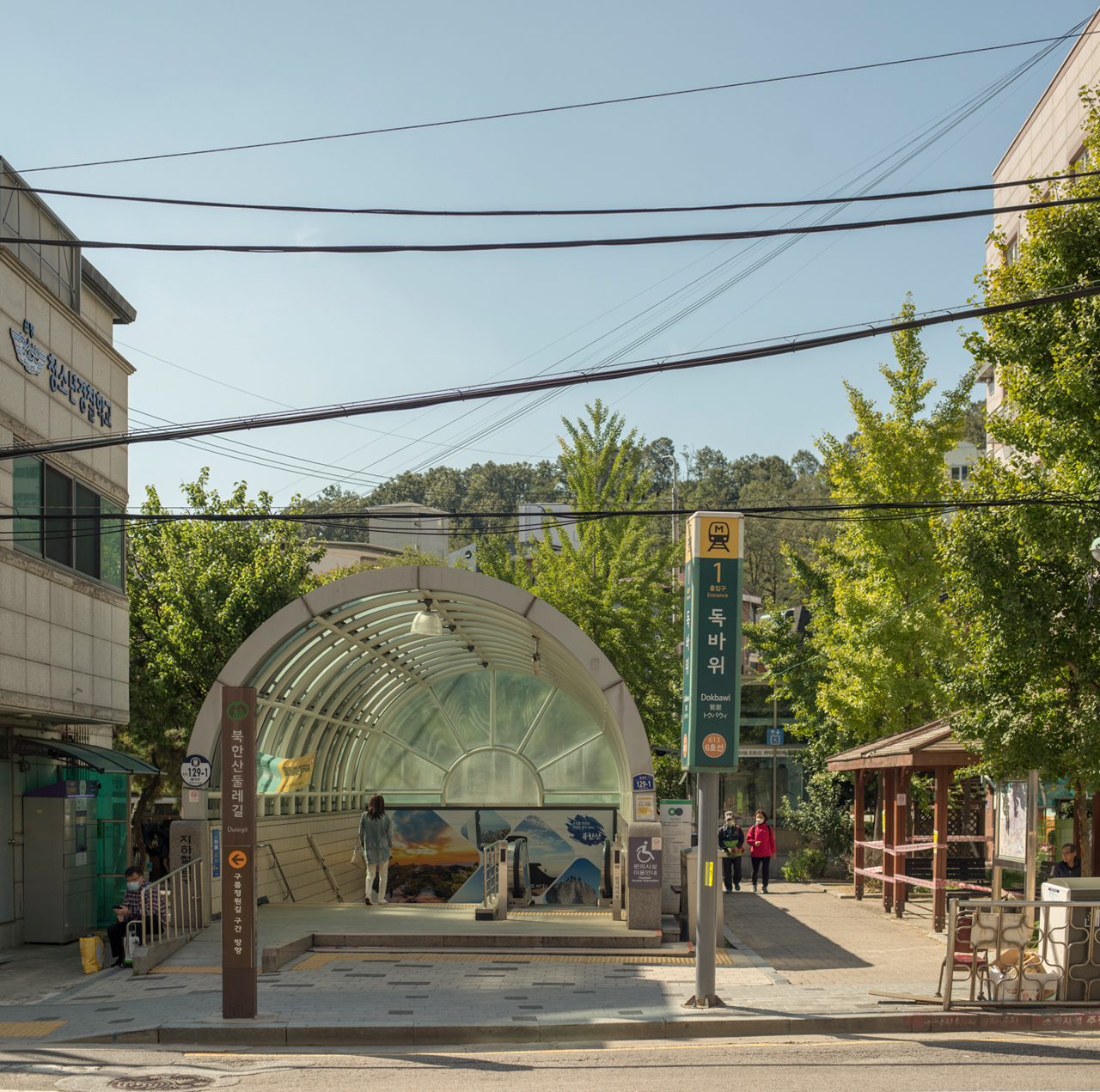The entrance of a subway on the edge of a wooded area with buildings and sidewalks and powerlines