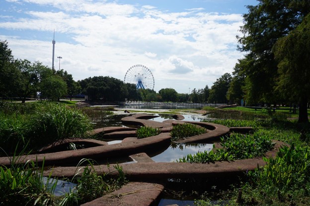 A picture of a body of water with twisting raised paths through it with trees and a Ferris wheel in the background