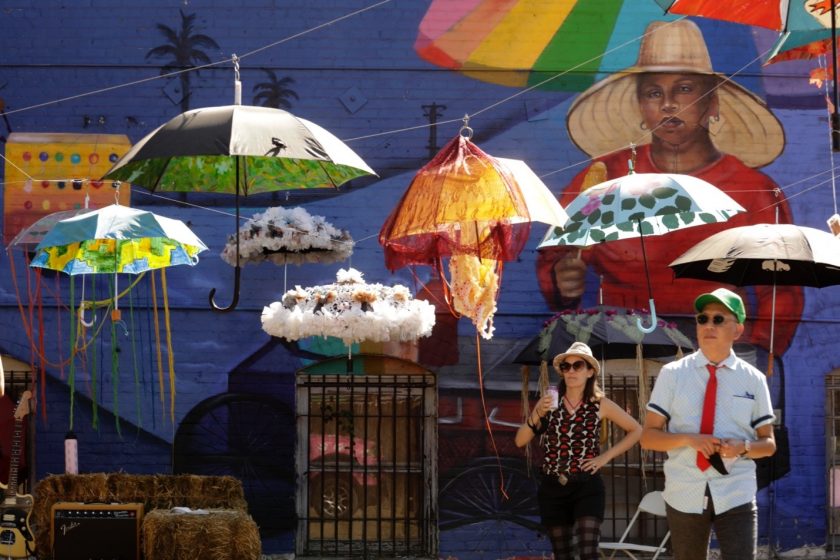 A picture of painted umbrellas hangings from strings in a courtyard with a mural in the background and people standing underneath