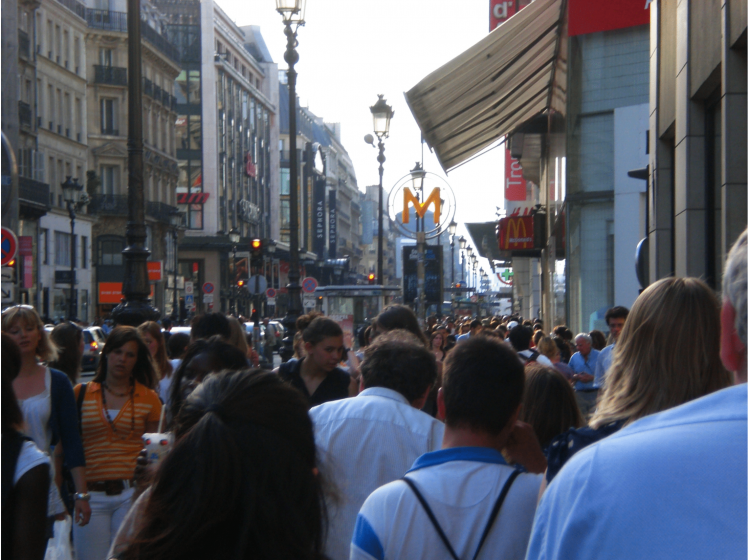 A picture of a crowd of people walking down a sidewalk along a street with buildings on both sides