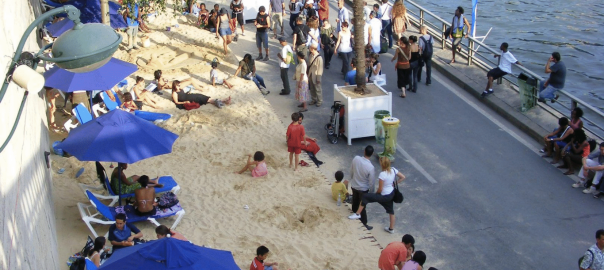 A picture of people relaxing on a small beach with blue umbrellas and a walkway next to the water