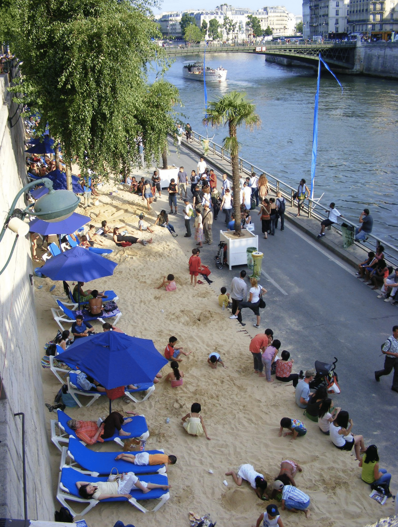 A picture of people relaxing on a small beach with blue umbrellas and a walkway next to the water