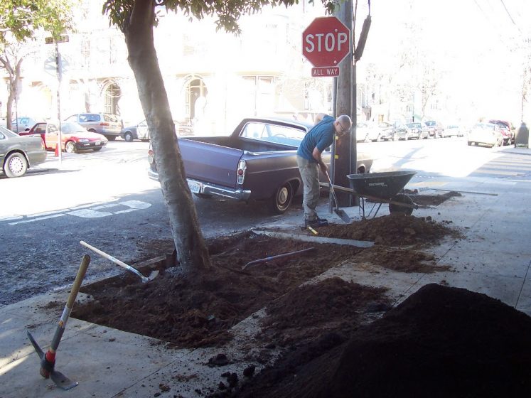 A picture of a man digging dirt on the side of a street with a car parked next to him