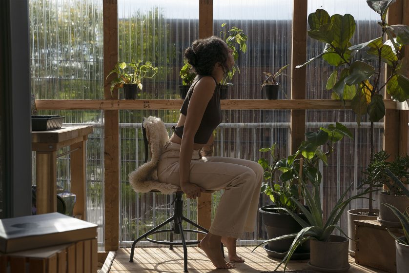 A picture of a woman sitting on a stool in a greenhouse looking out the window