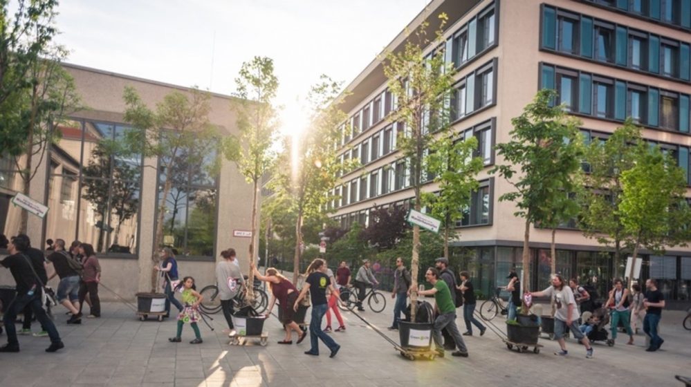 A picture of a sidewalk with people walking, cycling, and moving potted trees