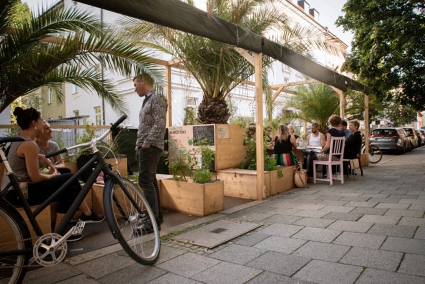 A picture of a sidewalk with outdoor seating with people sat and standing next to it