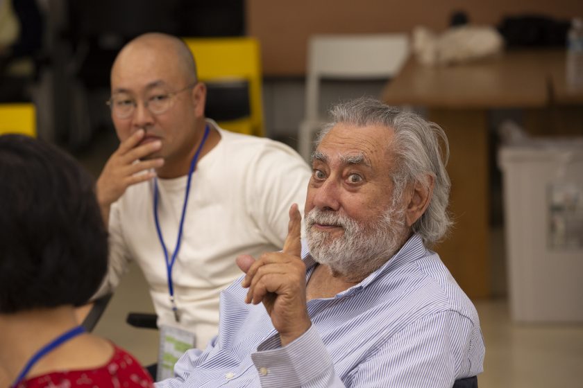 A picture of people sitting down at a conference talking with a man in center focus with his finger up