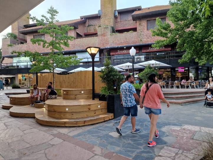 A picture of people walking next to a wooden sitting area with picnic tables, umbrellas, lights, and plants