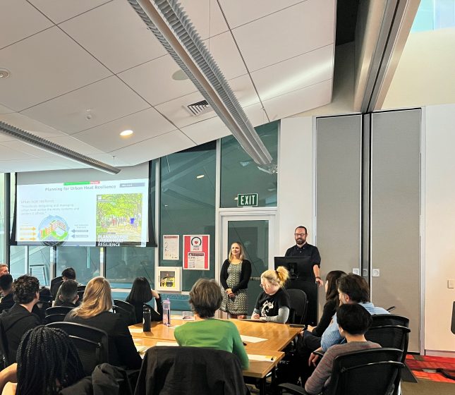 A picture of two people presenting a slide show in front of a group of people sitting around a table