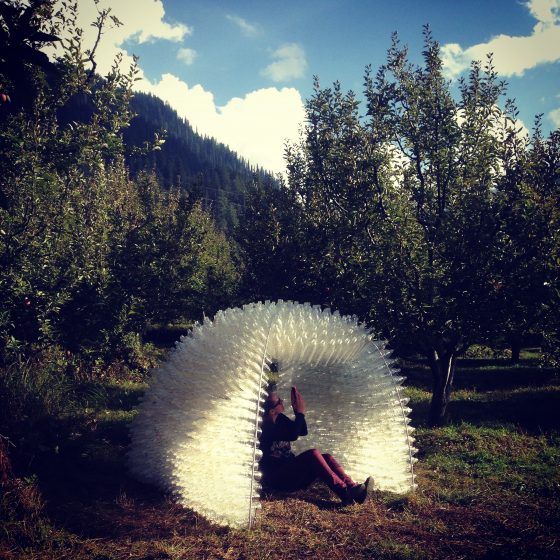 On a sunny day, a woman sits inside of a small structure made of plastic bottles, the structure surrounded by shrubs and trees in a mountain