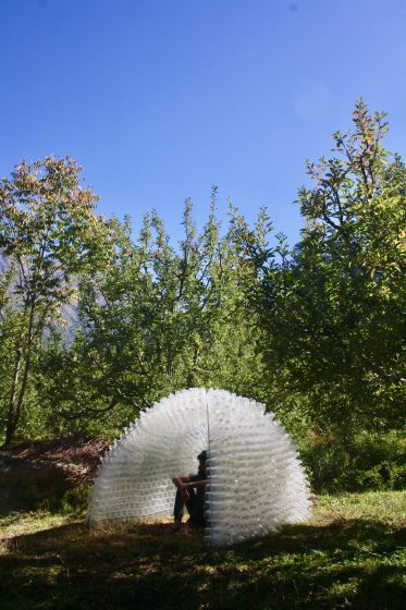 On a sunny day, a woman sits inside of a small structure made of plastic bottles, the structure surrounded by shrubs and trees