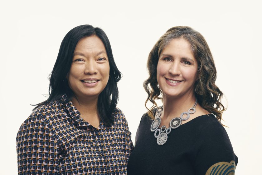 Two smiling women stand with each other in front of a white background.