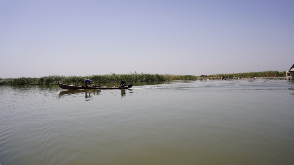 A longboat floats on murky water with a low-lying grass berm in the background.