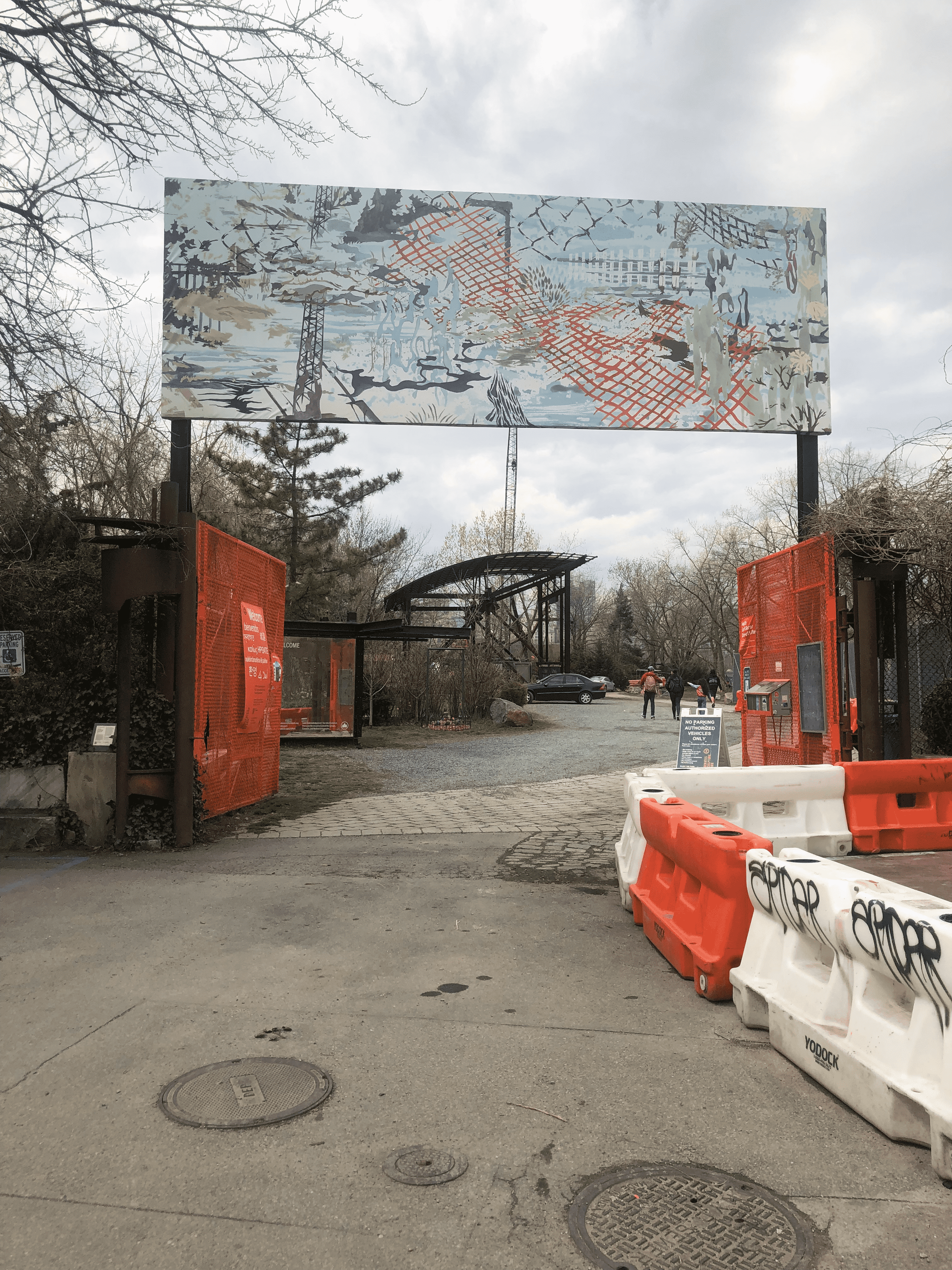 A picture of a road with orange and white barricades leading to an open orange gate with sculptures beyond it