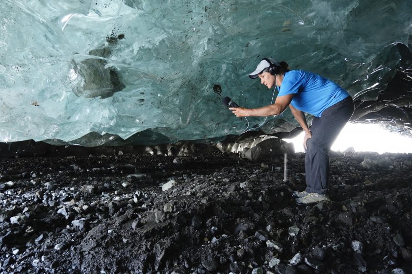 A woman with earphones and a microphone standing on rocky ground, under a large glistening blue wall of ice.