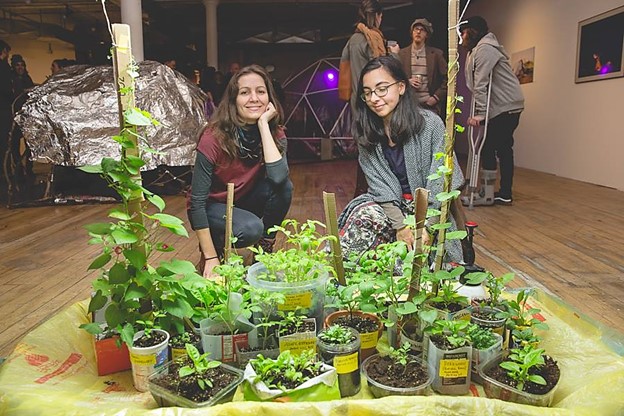 Two women posing behind a group of potted plants