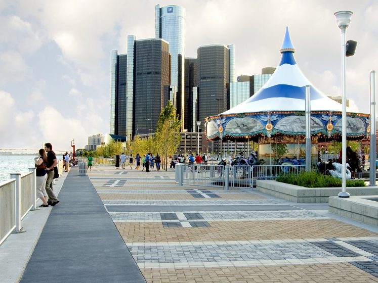 A group of people walking on a sidewalk next to a carousel with a city in the background