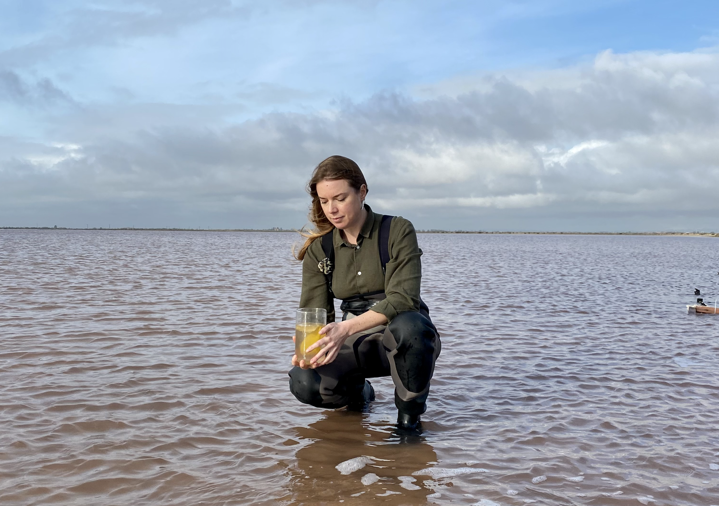 A person crouching in the water holding a beaker full of liquid