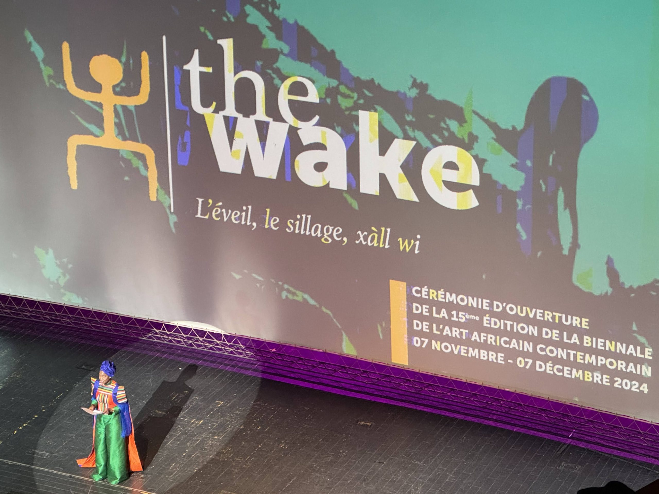 A person speaking on a stage with a giant sign behind them saying "the wake"