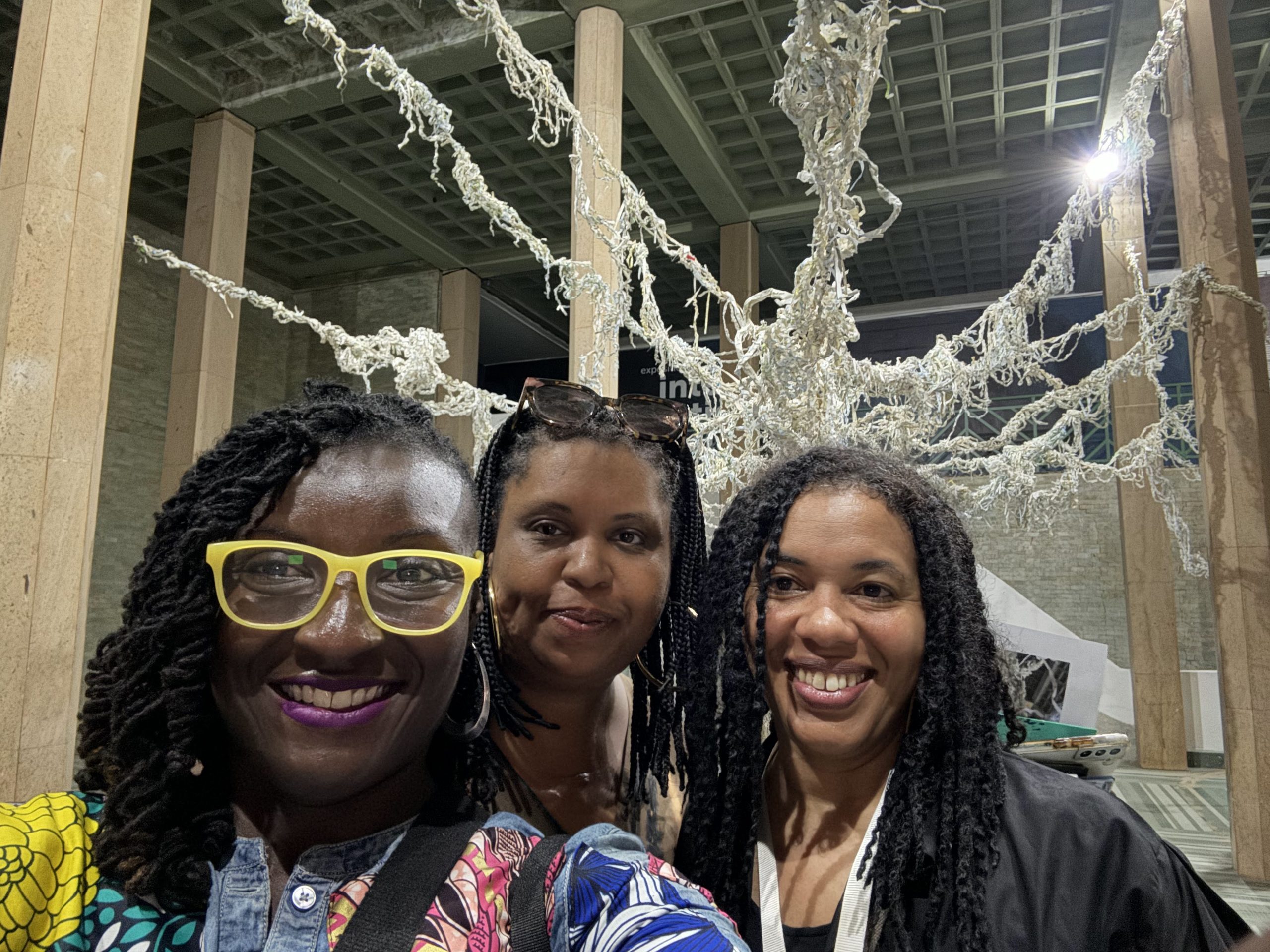 Three women posing in front of an art exhibit of shredded paper hanging from the ceiling in a web-like piece