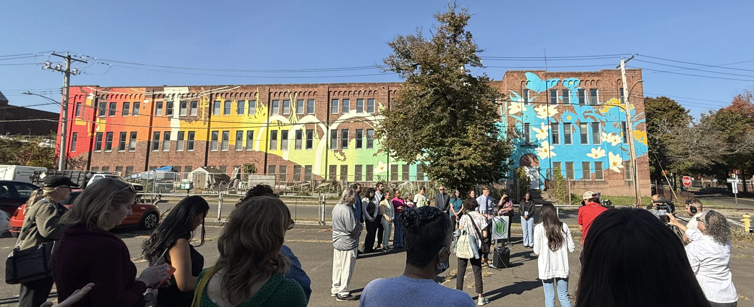A panoramic of people standing outside of a brick building looking at a mural