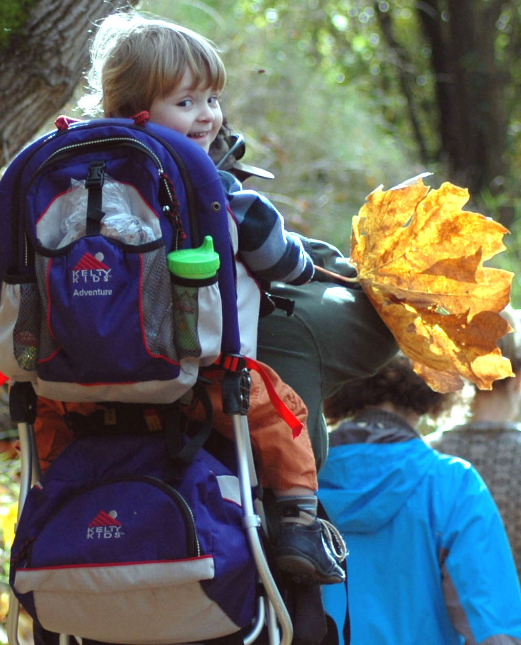 A young child in a backpack carrier smiling and holding a giant leaf