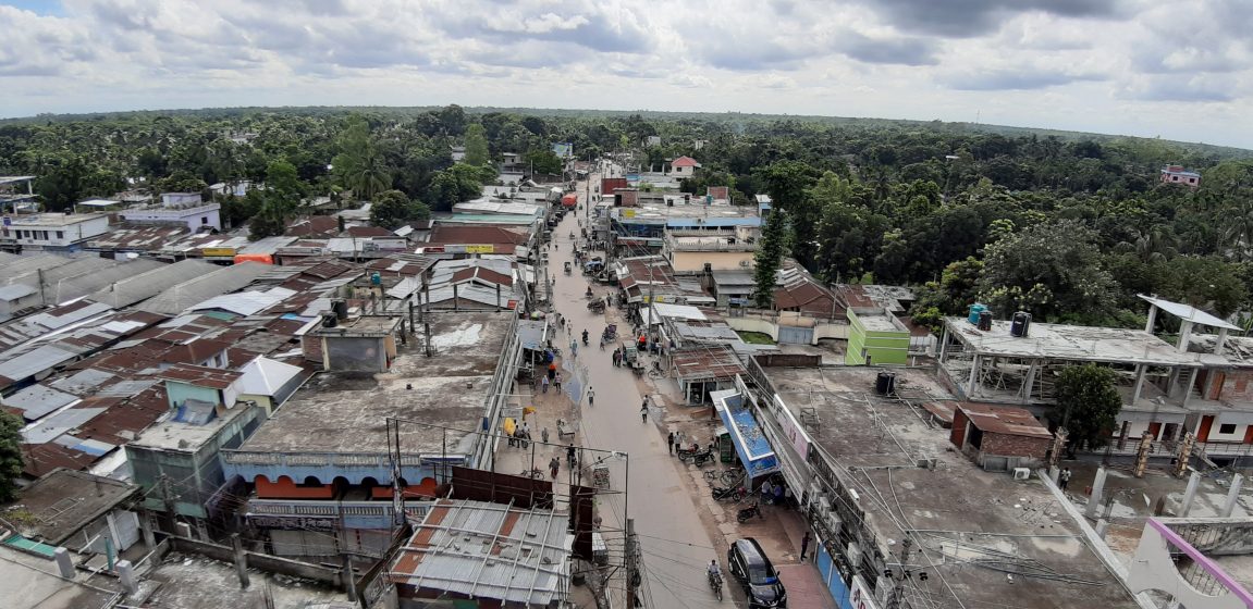 Aerial photograph of a busy street in a small town with numerous buildings and shops lining both sides. The scene includes people walking, vehicles parked and moving, and a backdrop of dense green trees under a cloudy sky.