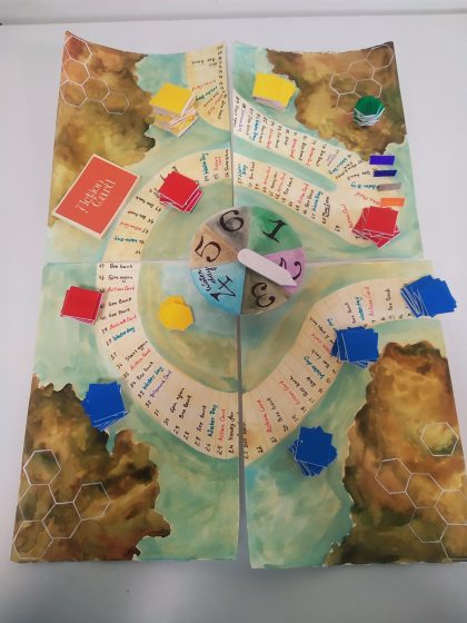 Board game layout featuring a hand-painted map with winding paths, hexagonal terrain sections, and a central spinner divided into numbered colored segments. Colored card stacks and tokens are placed along paths, indicating player positions and game progress on a fantasy-themed playing surface.