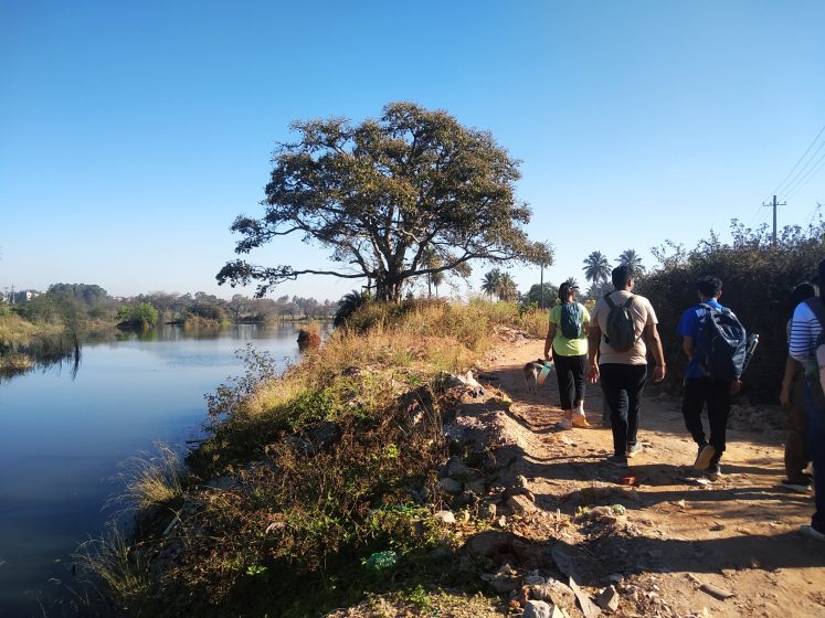 Photo of a group of people walking along a dirt path beside a calm river under a clear blue sky. The scene features a large tree near the riverbank, green vegetation, and power lines on the right.