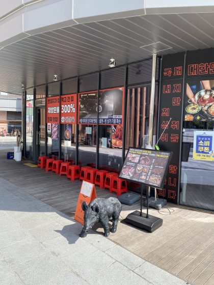 Photograph of a Korean barbecue restaurant exterior featuring large glass windows with red and black signage displaying menu items and promotional text. Red stools line the sidewalk seating area, a black pig statue stands near an orange sandwich board, and a menu stand with colorful food images is positioned at the entrance.