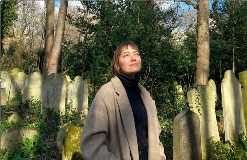 Photo of a person standing in an overgrown cemetery surrounded by old, weathered tombstones covered in moss and ivy. The scene features tall trees and dense greenery in the background, highlighting a quiet, natural setting with a mix of sunlight and shadows