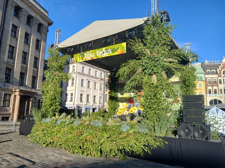 Outdoor stage decorated with abundant green plants and flowers, featuring a banner with partially visible text "Tirgus" under a clear blue sky. Surrounding historic buildings and large speaker stacks indicate a public event or market setting in an urban square.