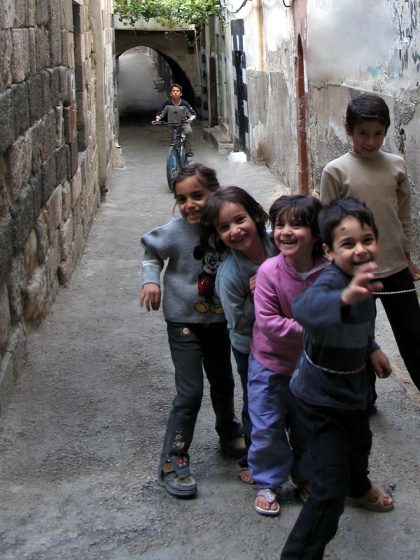 Photo of a narrow alleyway with five children standing in the foreground and one child riding a bicycle in the background. Walls on both sides show signs of wear and graffiti