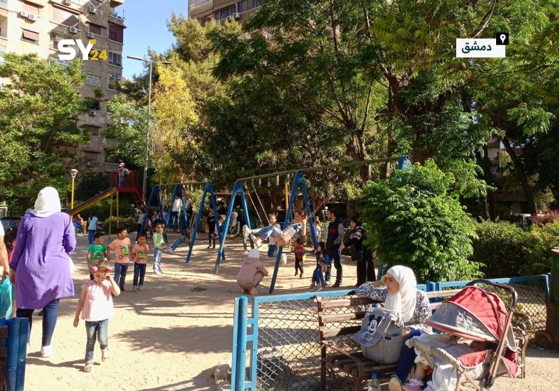 Photo of a busy playground in an urban park with children playing on swings and slides, surrounded by trees and apartment buildings. Bright daylight highlights colorful playground equipment, families, and caregivers