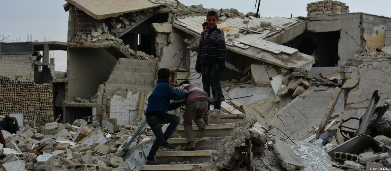 Photo showing three children navigating rubble and collapsed buildings in a war-torn urban area. The scene highlights extensive destruction with broken concrete, exposed rebar, and debris scattered around