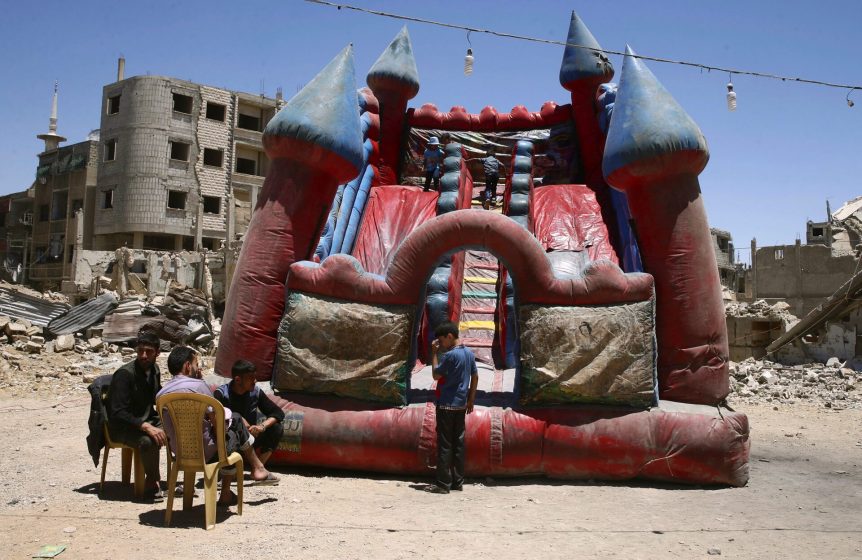 Photo showing a red and blue inflatable castle slide set up in a dusty, rubble-strewn area surrounded by damaged buildings. Four children sit on plastic chairs to the left while one child stands in front of the inflatable