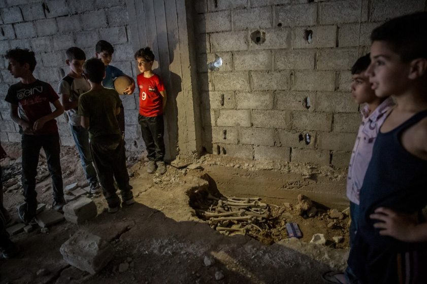 Photo showing a group of children standing around a partially unearthed human skeleton in a dimly lit, unfinished concrete room.