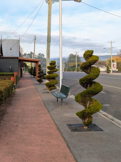 Photo of a sidewalk along a street featuring multiple spiral-shaped topiary trees evenly spaced near the curb. The scene includes a green bench, utility poles, and a building with a covered walkway, set against a backdrop of distant hills and a partly cloudy sky.
