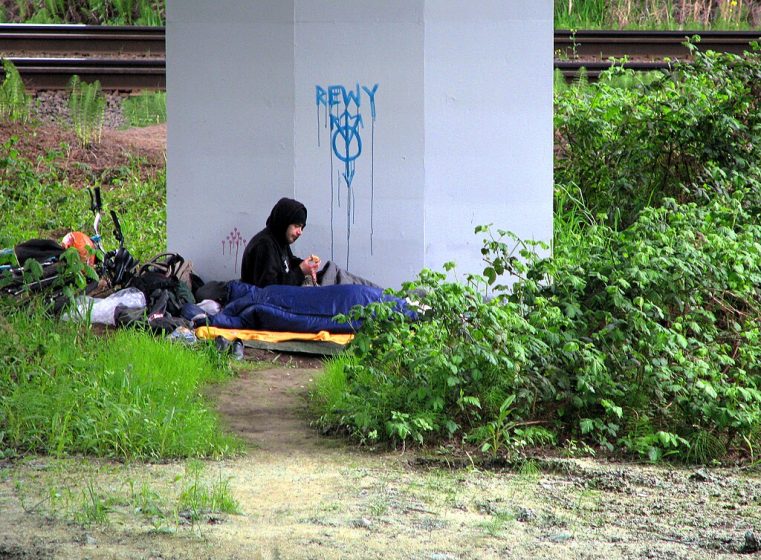 Photo of a person sitting under a bridge pillar near railroad tracks, surrounded by bags and blankets. Blue graffiti with the word "REWY" and a symbol is visible on the pillar, while green vegetation and dirt path frame the scene.