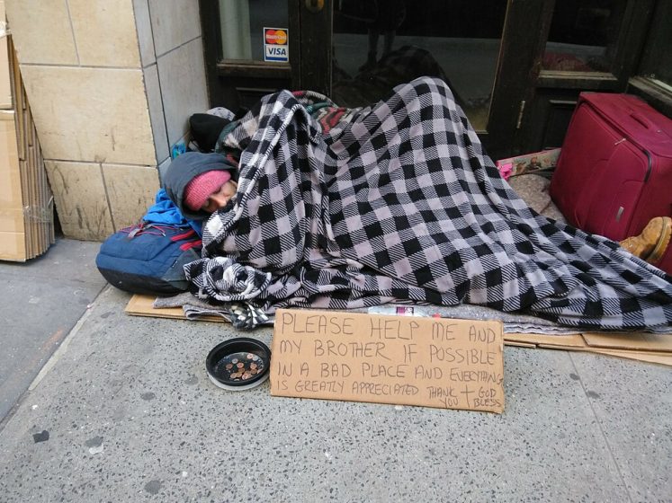 Photo of a person sleeping on a city sidewalk covered with a black and white checkered blanket, surrounded by personal belongings including a red suitcase and a blue backpack. A handwritten cardboard sign in front requests help for the person and their brother, mentioning they are in a bad place and expressing gratitude.
