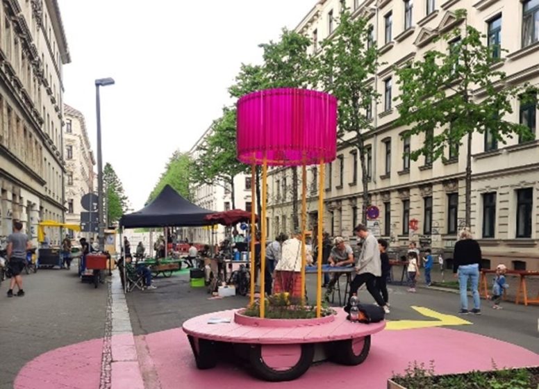 Photograph of a street scene featuring a circular pink-painted seating area with a tall, cylindrical magenta shade supported by yellow poles in the center. Surrounding the seating area are people walking and socializing, with buildings lining both sides of the street and a black tent visible in the background.
