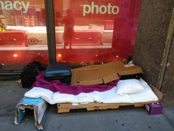 Photograph of a makeshift bed on a city sidewalk, constructed from cardboard, a wooden pallet, and various blankets and pillows. The setup is positioned against a building with red windows displaying partial words "pharmacy" and "photo". 