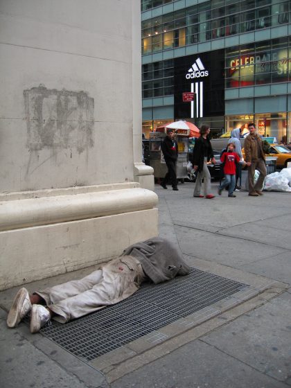 Photo of a person lying face down on a metal grate on a city sidewalk near a building corner. Background shows pedestrians walking past and a large Adidas store sign on a glass building across the street.