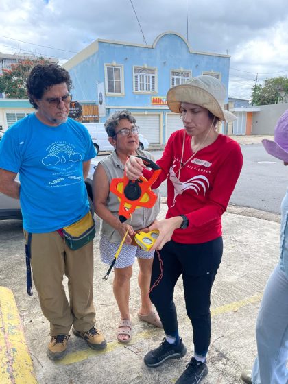 Photo of a person in a red shirt and sunhat demonstrating how to use a measuring device to a small group of people. They are in an urban setting with a light blue building in the background