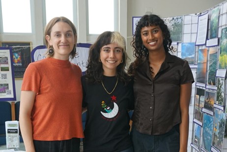 Photo of three people standing indoors in front of a display board with various printed photographs of nature scenes. The individuals wear casual clothing in orange, black, and dark brown, with natural light coming from windows behind them.