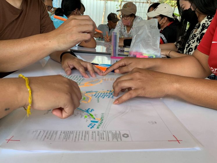 Photo of a group of people collaborating around a table, using a transparent ruler and colorful markers to work on a detailed document or map. Focus is on hands actively measuring and marking, with various tools and materials visible.