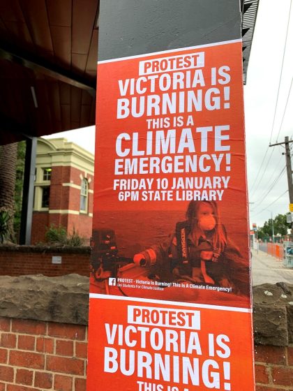Poster on pole announces a protest event titled "Victoria is Burning! This is a Climate Emergency" scheduled for Friday, January 10 at 6 PM at State Library. The poster features bold white text on a red background with an image of a firefighter in protective gear and includes social media handles and a call to action for climate emergency awareness.