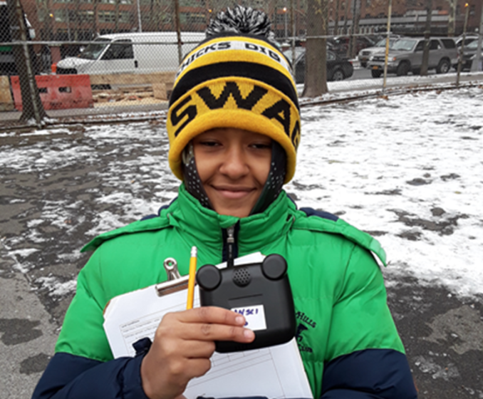 Photo of a person outdoors in winter clothing holding a clipboard with papers and a small black device labeled "WSI." Background shows a snowy ground, chain-link fence, and parked vehicles,