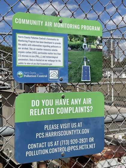 Photo of a community air monitoring program sign attached to a chain-link fence, featuring green and blue panels with white text. The sign explains Harris County Pollution Control's initiative to monitor air pollutants like VOCs and PM2.5, provides a website for data access, and lists contact information for reporting air-related complaints.